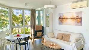 A modern living room with a sofa, coffee table, dining table, and chairs. There is a painting on the wall and an air conditioning unit above the sofa. - Apartment on the Beach with Sea Views, Balcony, Shared Pool & WiFi (Nerja)