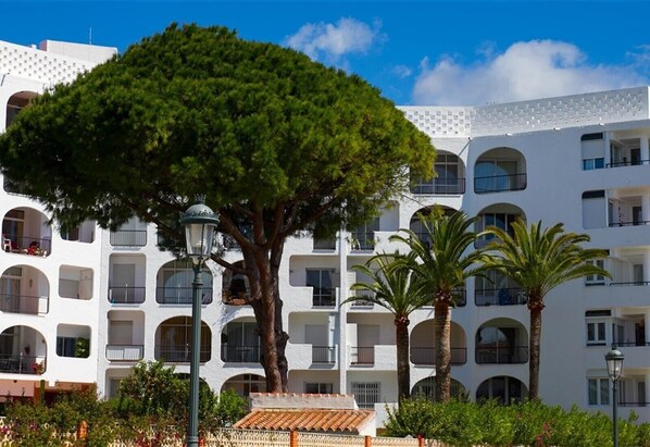 Un edificio a più piani con balconi, un albero grande e palme.