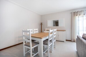 A small, clean room with a wooden dining table, white chairs, a white cabinet, a mirror, and a grey sofa.