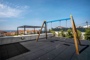 A rooftop with a swing set, a glass enclosure, and a view of buildings and the sea.