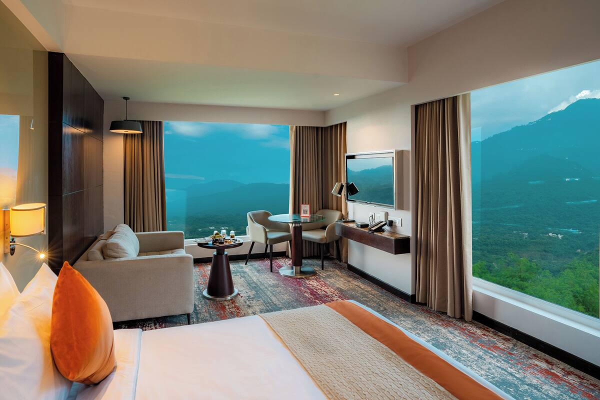 Quarto de hotel com cama, sofá, escrivaninha e cadeira. Há uma janela grande com vista para as montanhas.