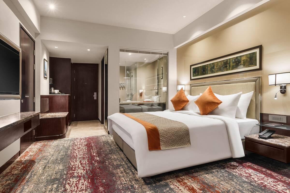 Quarto de hotel com uma cama grande, uma escrivaninha, um espelho e um banheiro visível através de uma porta aberta.