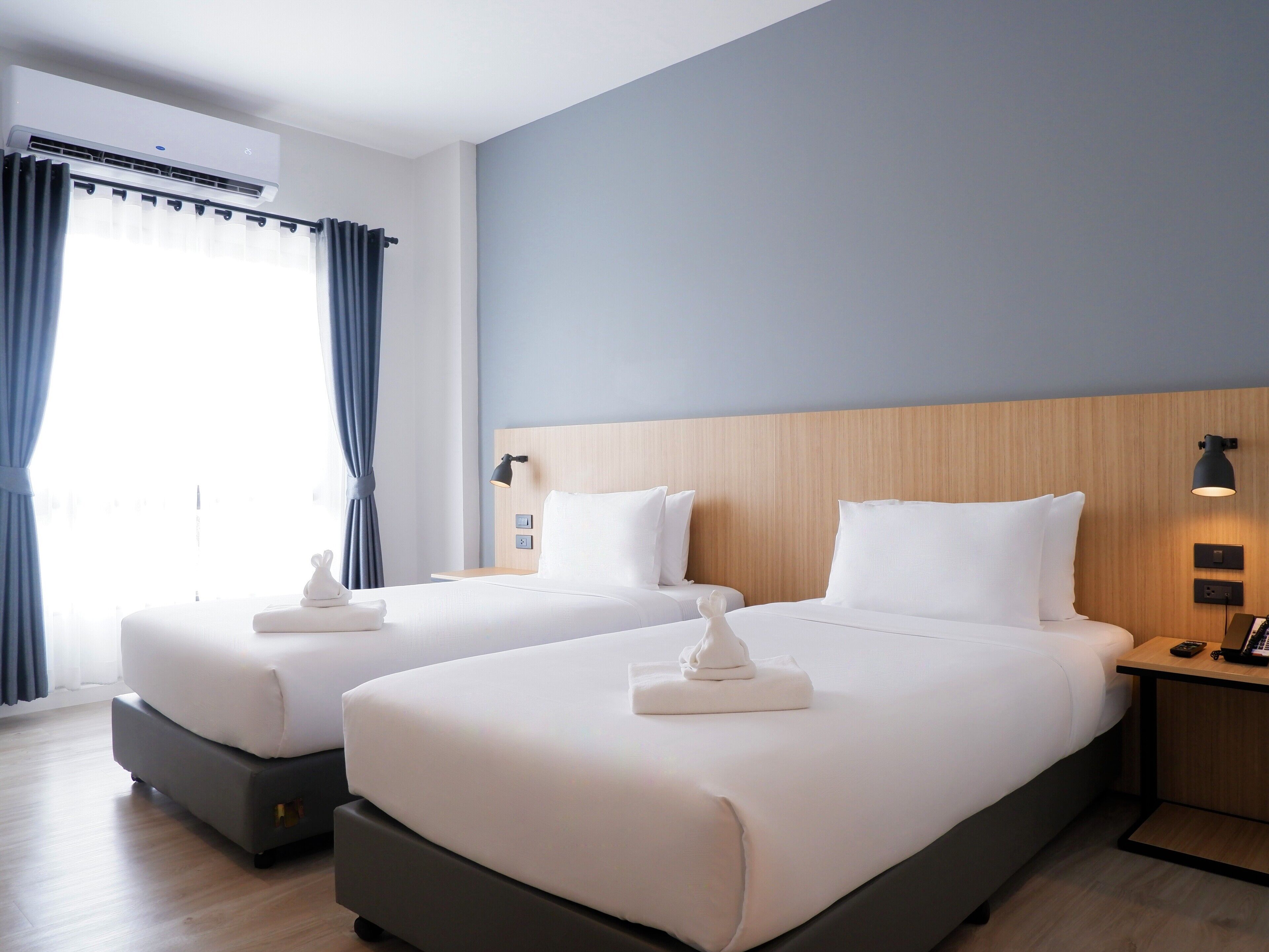 A hotel room with two beds, a wooden headboard, a bedside table with a lamp, and a window with curtains.