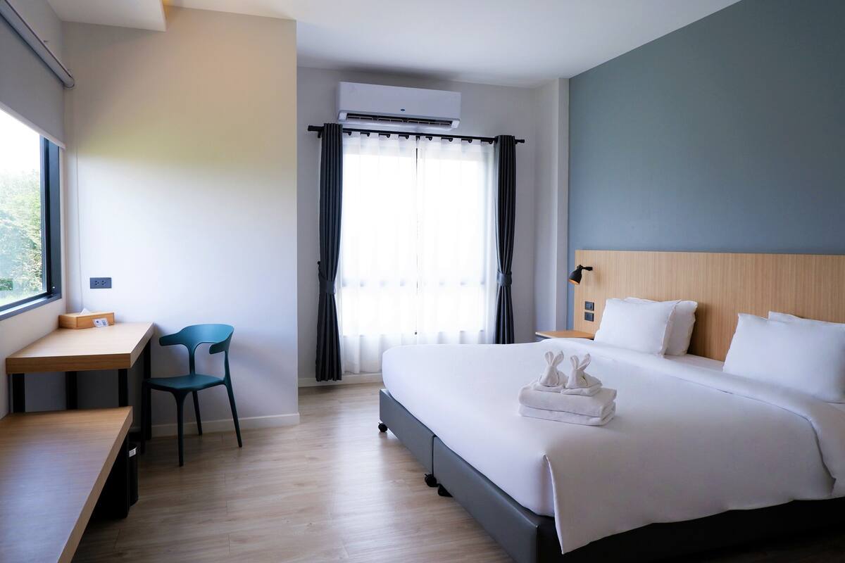 A hotel room with a bed, a desk, a chair, and an air conditioning unit.