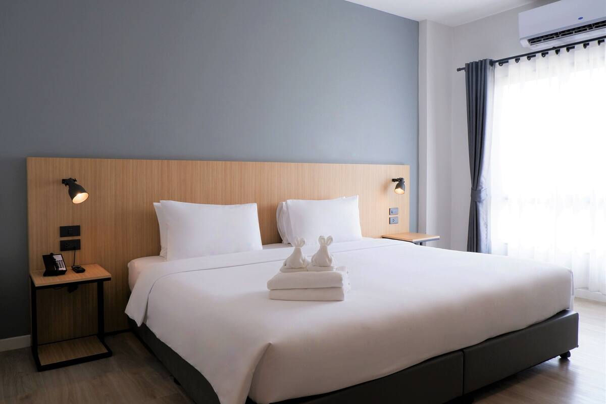A modern hotel room with a wooden headboard, a large bed with white linens, two bedside tables with lamps, and a view of the city through sheer curtains.