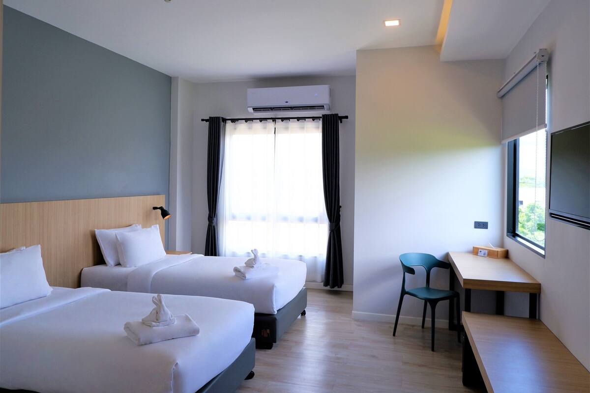 A hotel room with two beds, a desk, a chair, an air conditioning unit, and a window with curtains.