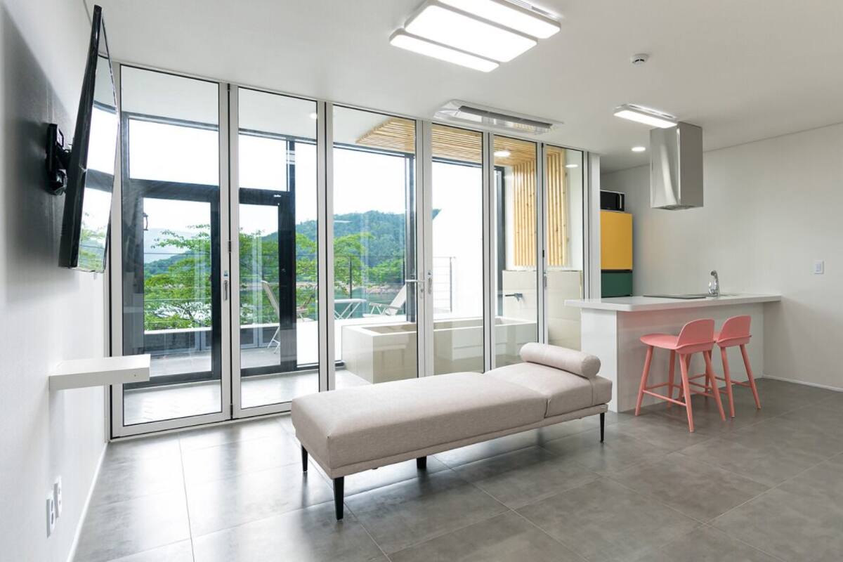 A modern living room with a sofa, a dining area with pink chairs, and a large window offering a view of greenery.