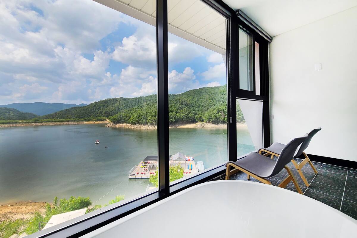 A room with a large window overlooking a lake, a chair, and a bathtub.