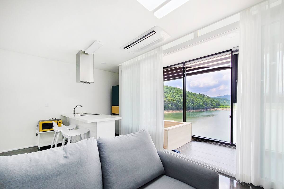 A modern living room with a grey sofa, a white desk with a chair, a small table, and a large window offering a view of a lake and greenery.