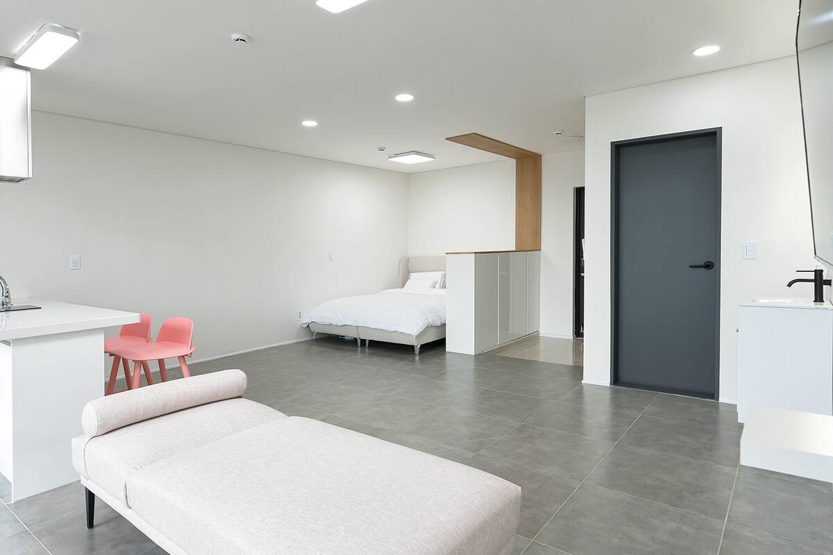A modern hotel room with a kitchenette, a bed, a desk with a chair, and a TV mounted on the wall.