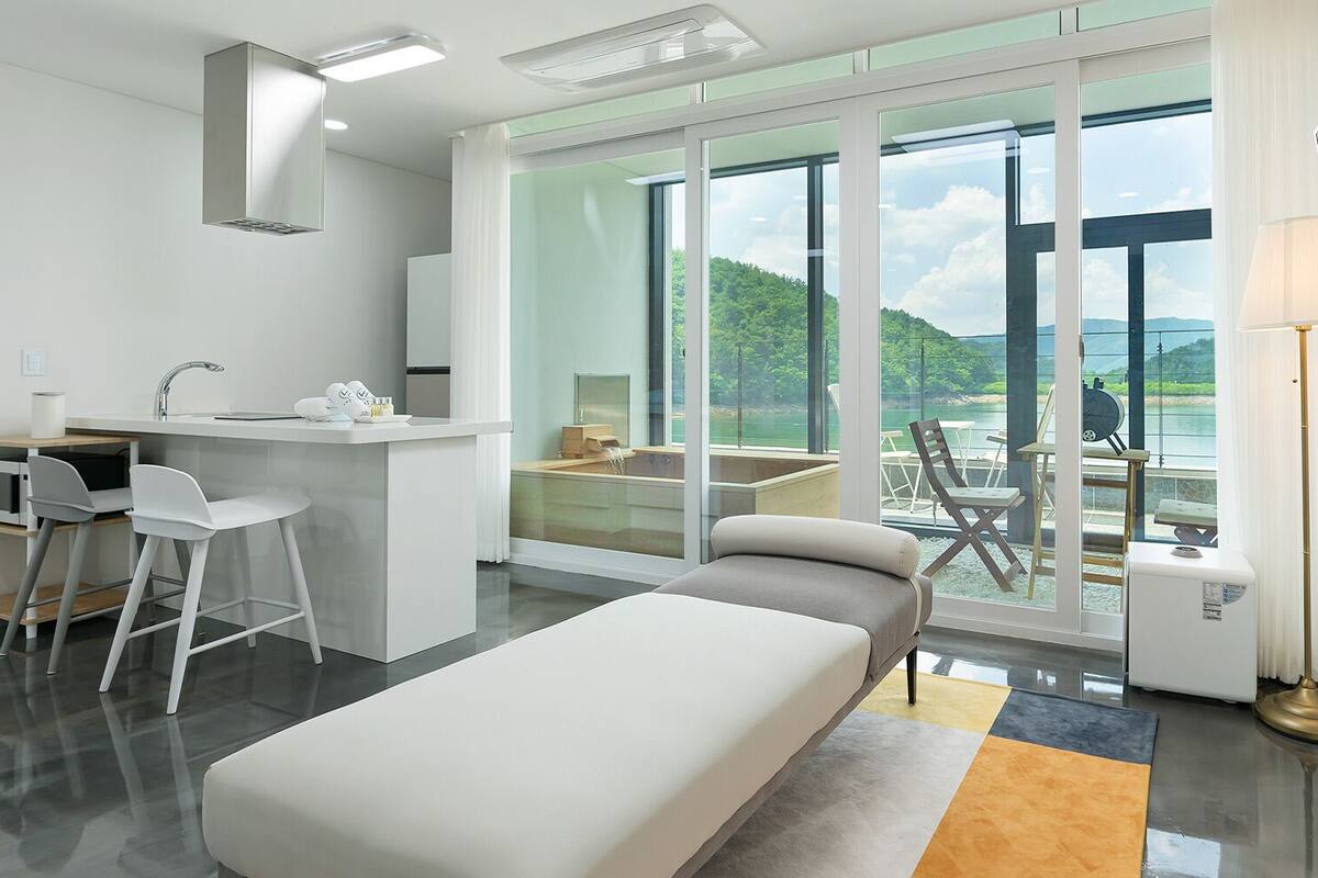 A modern hotel room with a bed, a desk with chairs, a large window with a view of a lake and mountains, and a balcony with outdoor seating.