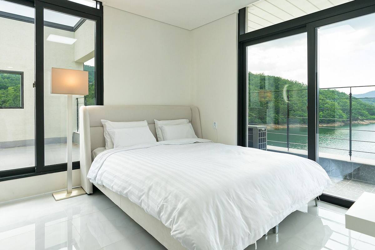 A modern bedroom with a large bed, a nightstand, a lamp, and a view of a lake and mountains through large windows.