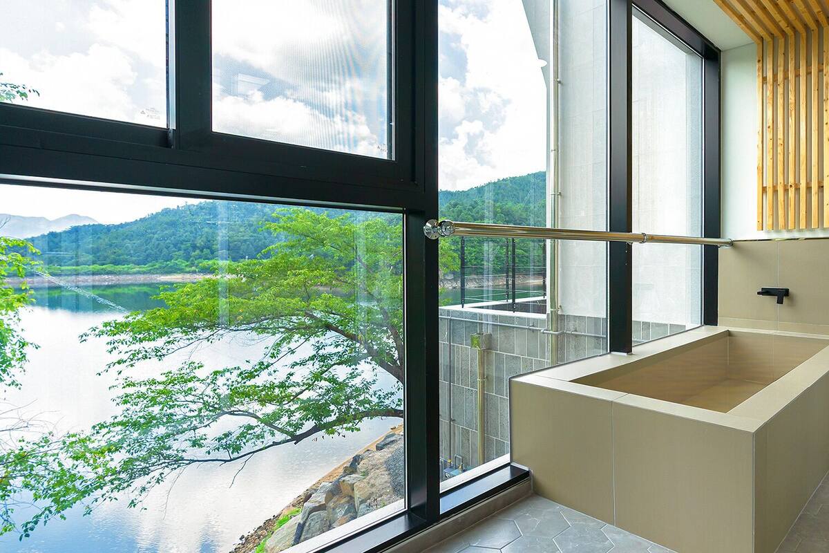 A modern bathroom with a large window, a bathtub, and a view of a river and mountains.
