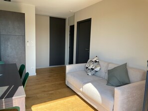 A modern living room with a sofa, a dining table, and chairs. There is a hallway leading to a door.