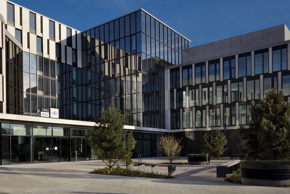A modern building with a glass facade, a clear sky, and a landscaped courtyard with trees and seating areas. - Holiday Inn - the niu, Hub Dusseldorf Messe by IHG (Düsseldorf)