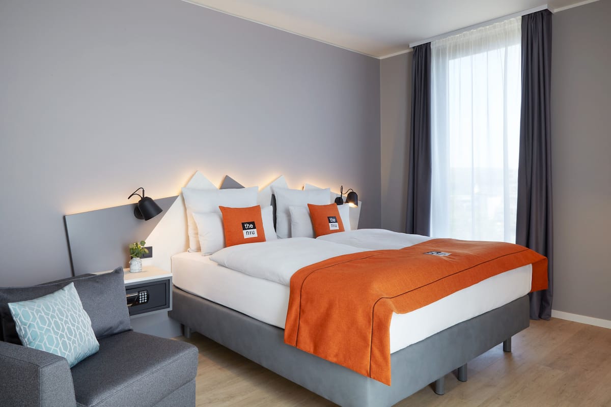 A modern bedroom with a large bed, grey and white bedding, a grey sofa, a bedside table with a lamp, and a window with curtains.
