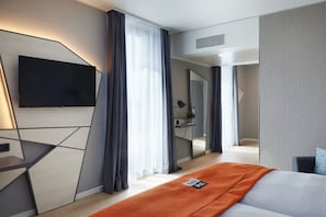 Una habitación de hotel moderna con una televisión de pantalla plana montada en la pared, una cama con una manta naranja y un espejo grande que refleja la habitación.