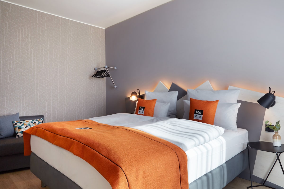 A modern hotel room with a neatly made bed, a side table with a lamp, and a small plant.