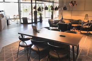 Una moderna hall d'albergo con una lunga reception, diverse aree salotto e ampie finestre che lasciano entrare la luce naturale.