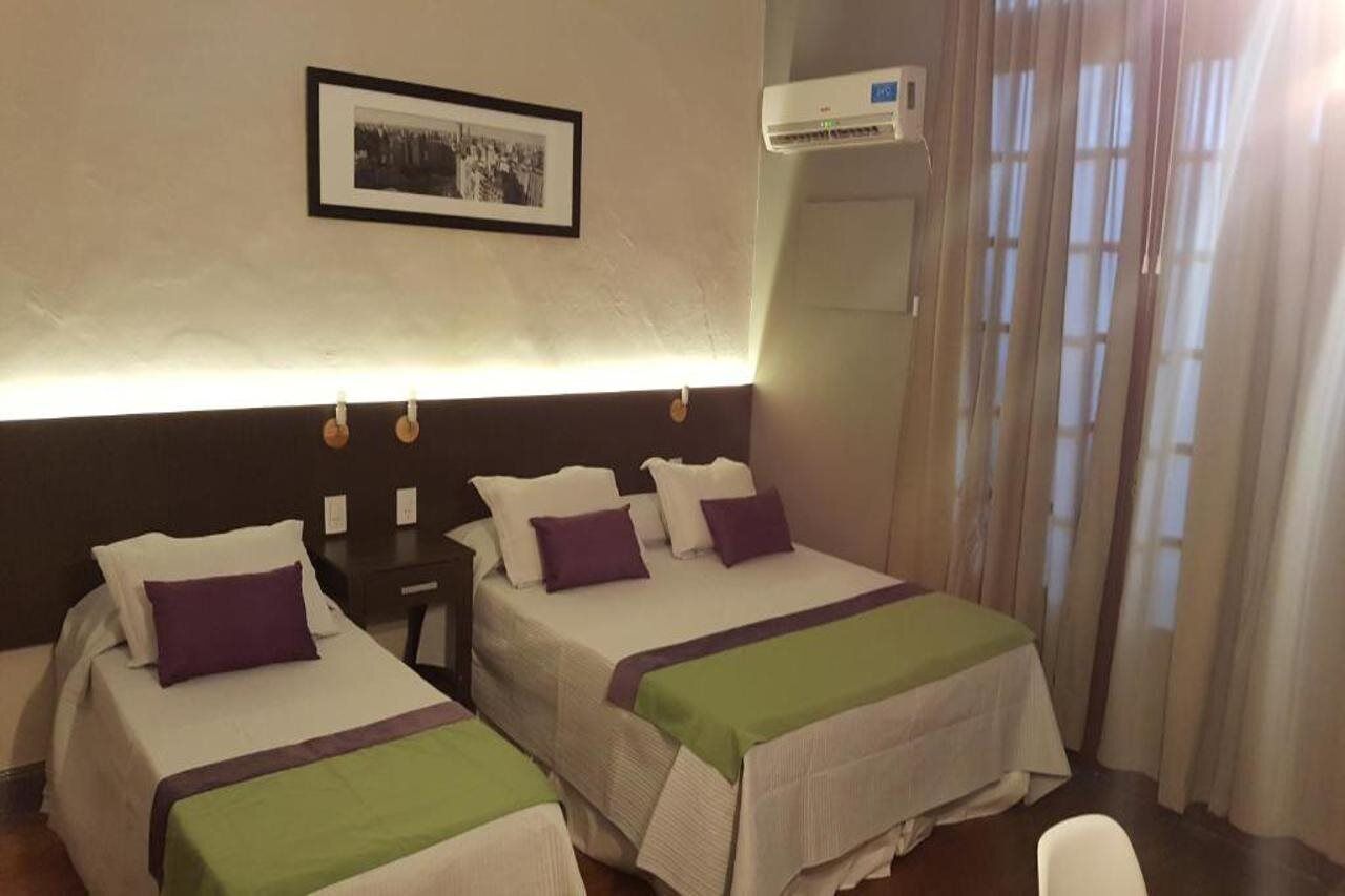 A hotel room with two beds, a bedside table, a framed picture on the wall, and an air conditioning unit.