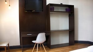 Habitación compacta con cama, escritorio con silla, televisor montado en la pared y estante con cestas.