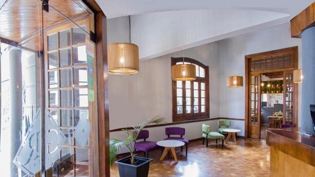 Un vestíbulo de hotel con un mostrador de recepción de madera, varias zonas de estar con sillas y mesas, y amplios ventanales que permiten la entrada de luz natural.