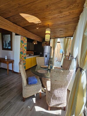 Relax in this inviting room with a round table and wicker chairs, a modern kitchen, and a large window with sheer curtains.