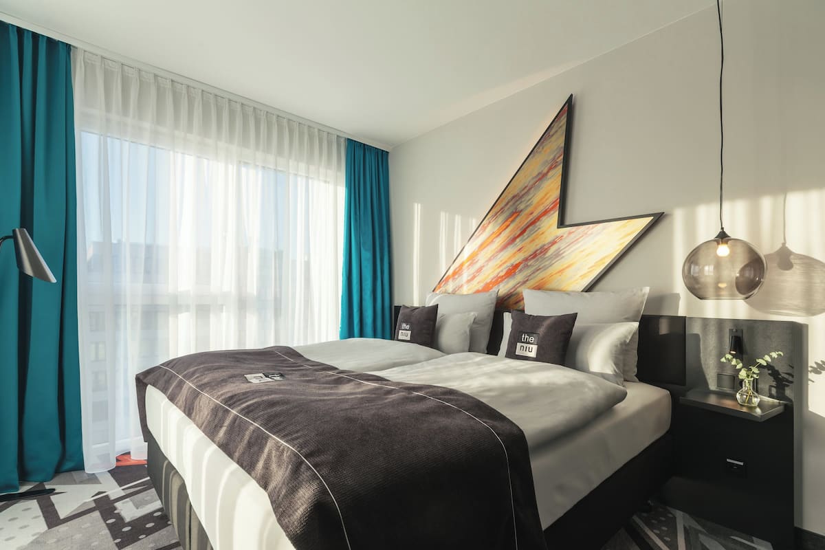 A modern hotel room with a large bed, a bedside table with a lamp, and a large window with curtains.