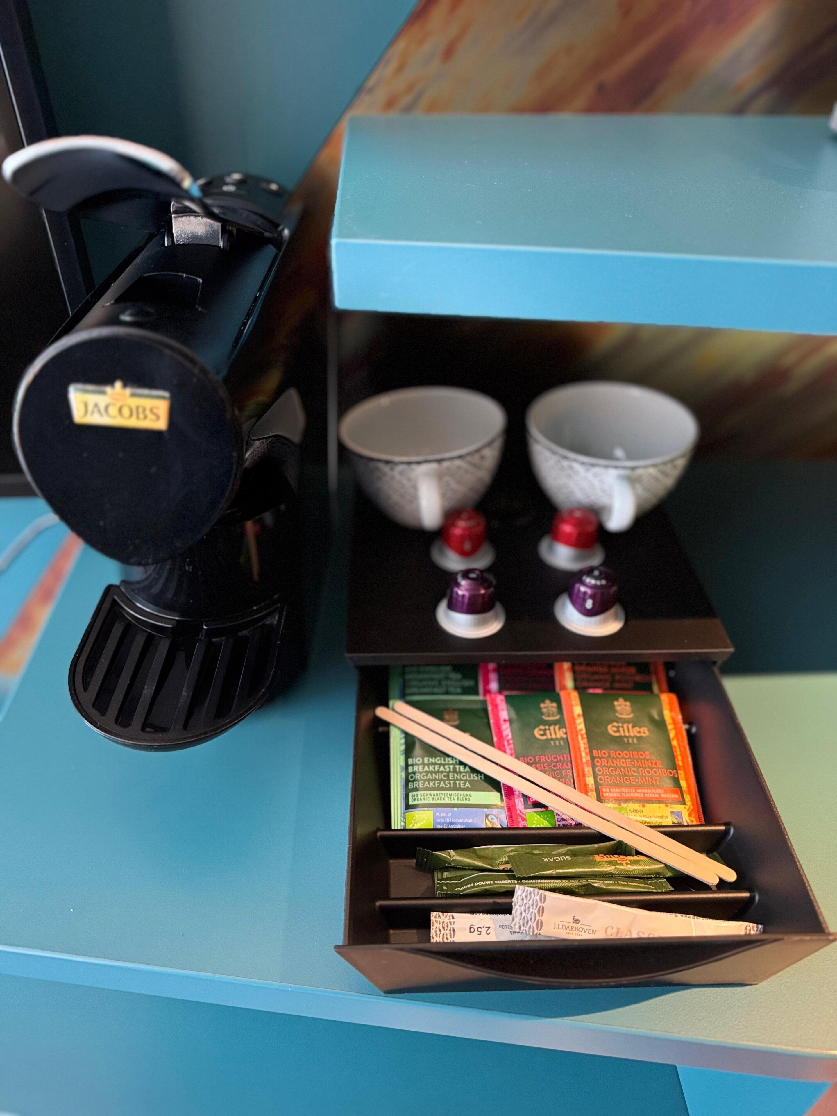 Une machine à café de la marque Jacobs, deux tasses et un plateau contenant des sachets de thé et des baguettes.