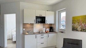 Ett modernt kök med vita skåp, en svart mikrovågsugn och en kaffebryggare. Det finns ett fönster med utsikt över ett hus och en tavla med blommor på väggen.