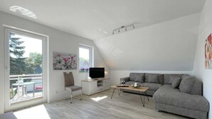 Ett modernt vardagsrum med en grå soffa, en tv på ett träställ och en balkong med utsikt över träd och ett hus.