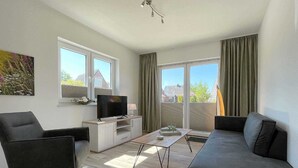 A modern living room with a sofa, a TV on a stand, a coffee table, and a balcony with curtains.