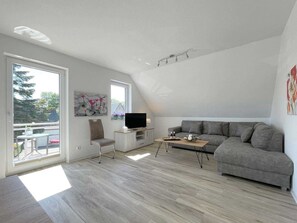 Uma sala de estar moderna com um sofá cinzento, uma TV sobre um móvel de madeira e uma varanda com vista para árvores e uma casa.