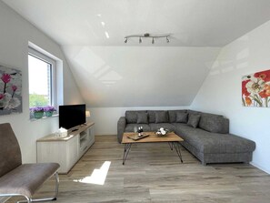 Ein modernes Wohnzimmer mit einem grauen Ecksofa, einem Holztisch, einem Fernseher auf einem Holzständer und einem Fenster mit Blumentöpfen.