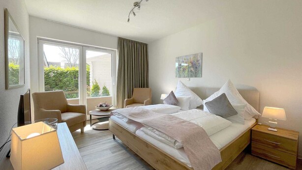 Ein Hotelzimmer mit einem großen Bett, zwei Nachttischen, einem Schreibtisch mit Stuhl, einem zusätzlichen Sessel, einem kleinen Tisch mit Obstschale und einem Fenster mit Vorhängen.