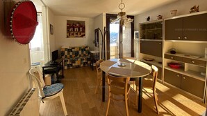 Un coin repas avec une table ronde, des chaises, un chapeau rouge accroché au mur, un tableau et une vue sur l’extérieur, visible à travers la porte.