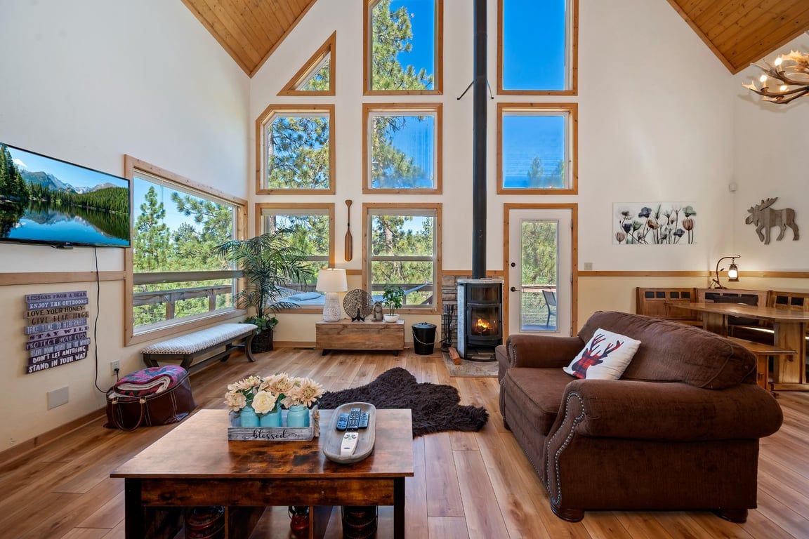 A spacious living room with a large TV, wooden furniture, a fireplace, and large windows offering a view of the outdoors.