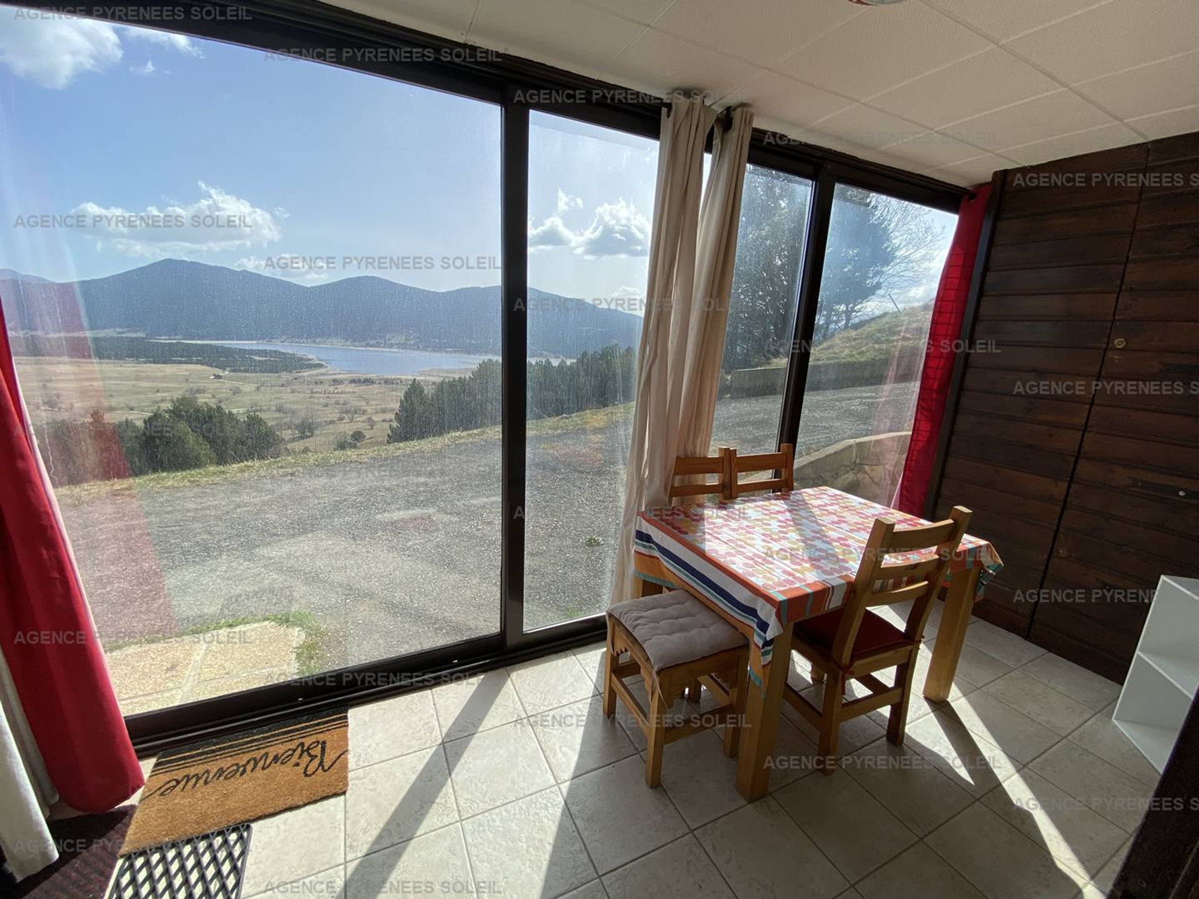 Una habitación con un ventanal que ofrece vistas a un lago y montañas. Hay una mesa de madera con sillas y una alfombra pequeña.