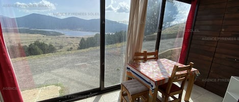 Una habitación con un ventanal que ofrece vistas a un lago y montañas. Hay una mesa de madera con sillas y una alfombra pequeña.