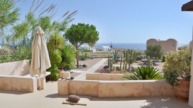 Une terrasse avec un canapé beige, une table et un pot de fleurs. On aperçoit la mer et un bâtiment au loin.
