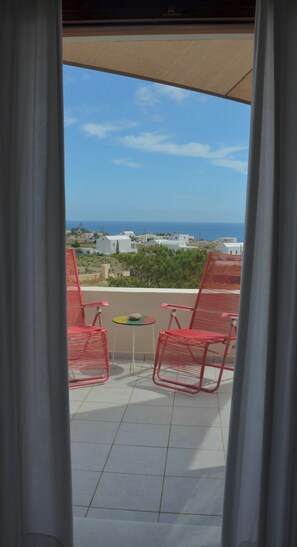 A balcony with two red chairs and a small table, overlooking a coastal area with buildings and the sea.