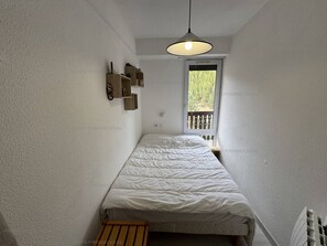 A narrow room with a bed, a wooden bench, a hanging light fixture, and a window with a view of greenery. - Cozy Ski-in/Ski-out Apartment with Balcony, WiFi & Parking - Sleeps 6 (Les Angles)