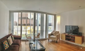Ein modernes Wohnzimmer mit einer Couch, einem Glastisch, einer TV-Konsole und einem Balkon mit großen Fenstern.