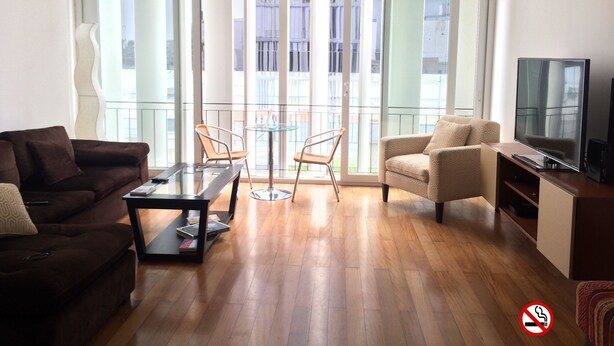 A living room with a brown sofa, a glass coffee table, a beige armchair, a TV, and a balcony with a glass railing. - Stunningly Beautiful & Modern Apt. Small building (Miraflores)
