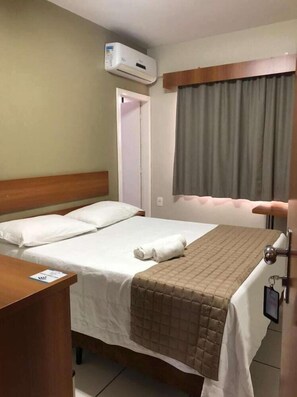 A hotel room with a bed, a bedside table, an air conditioning unit, a window with curtains, and a door. - Tri Hotel Centro Chapeco (Chapecó)