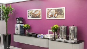 A reception area with a purple wall, a coffee machine, two framed pictures, a potted plant, and a coffee dispenser. - Tri Hotel Centro Chapeco (Chapecó)