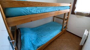 Een stapelbed met blauw beddengoed in een kleine kamer met een raam en een radiator.