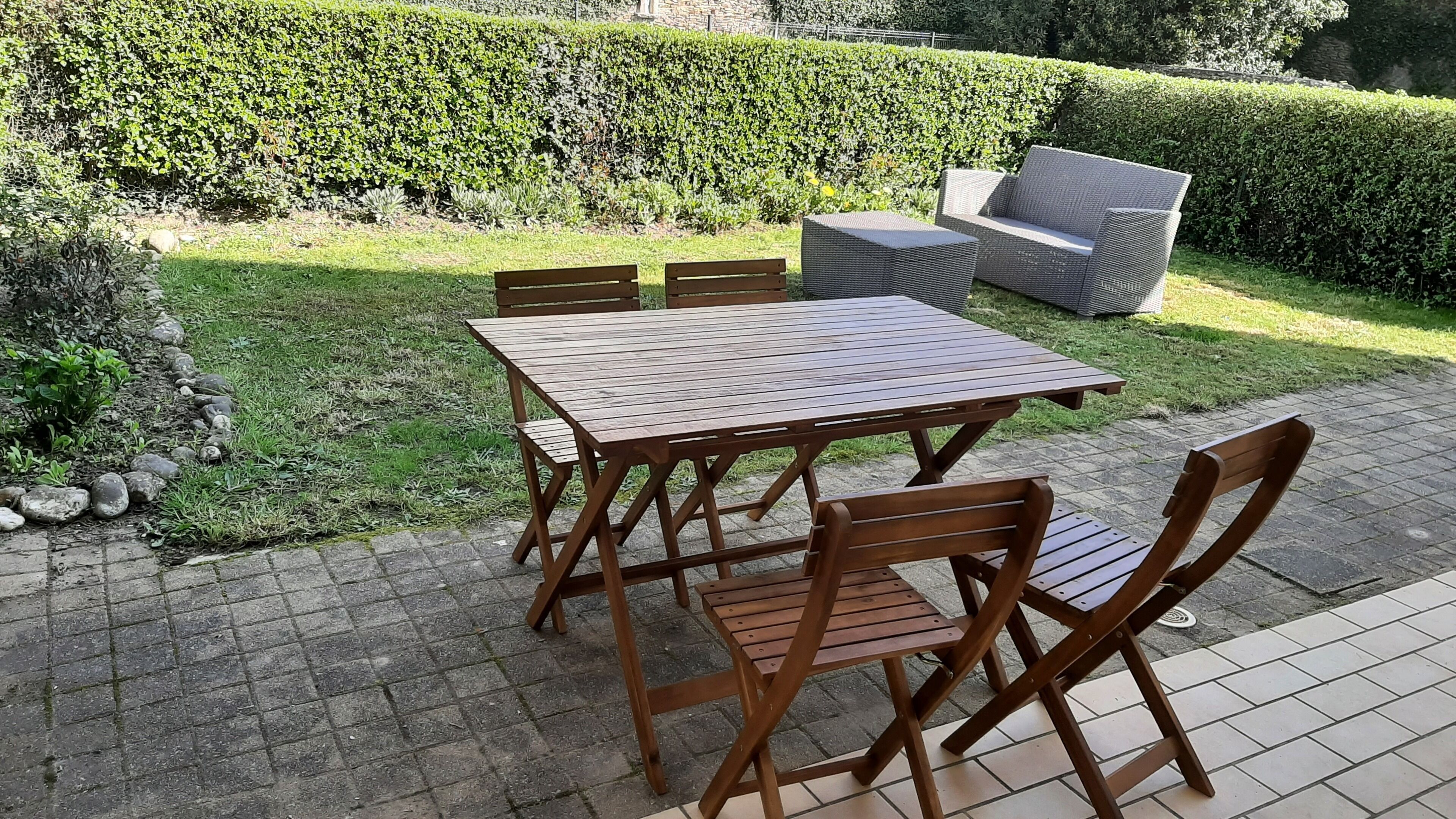 A patio area with a wooden table and chairs, a cushioned sofa, and a well-maintained hedge in the background.