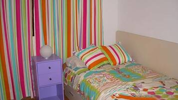 Een slaapkamer met een bed, kleurrijke gestreepte gordijnen, een klein paars nachtkastje met lades en een witte muur.