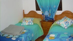 Een kamer met twee eenpersoonsbedden, elk voorzien van een beddekleed met een patroon en passende kussens. Daartussen staat een houten nachtkastje met een lamp.
